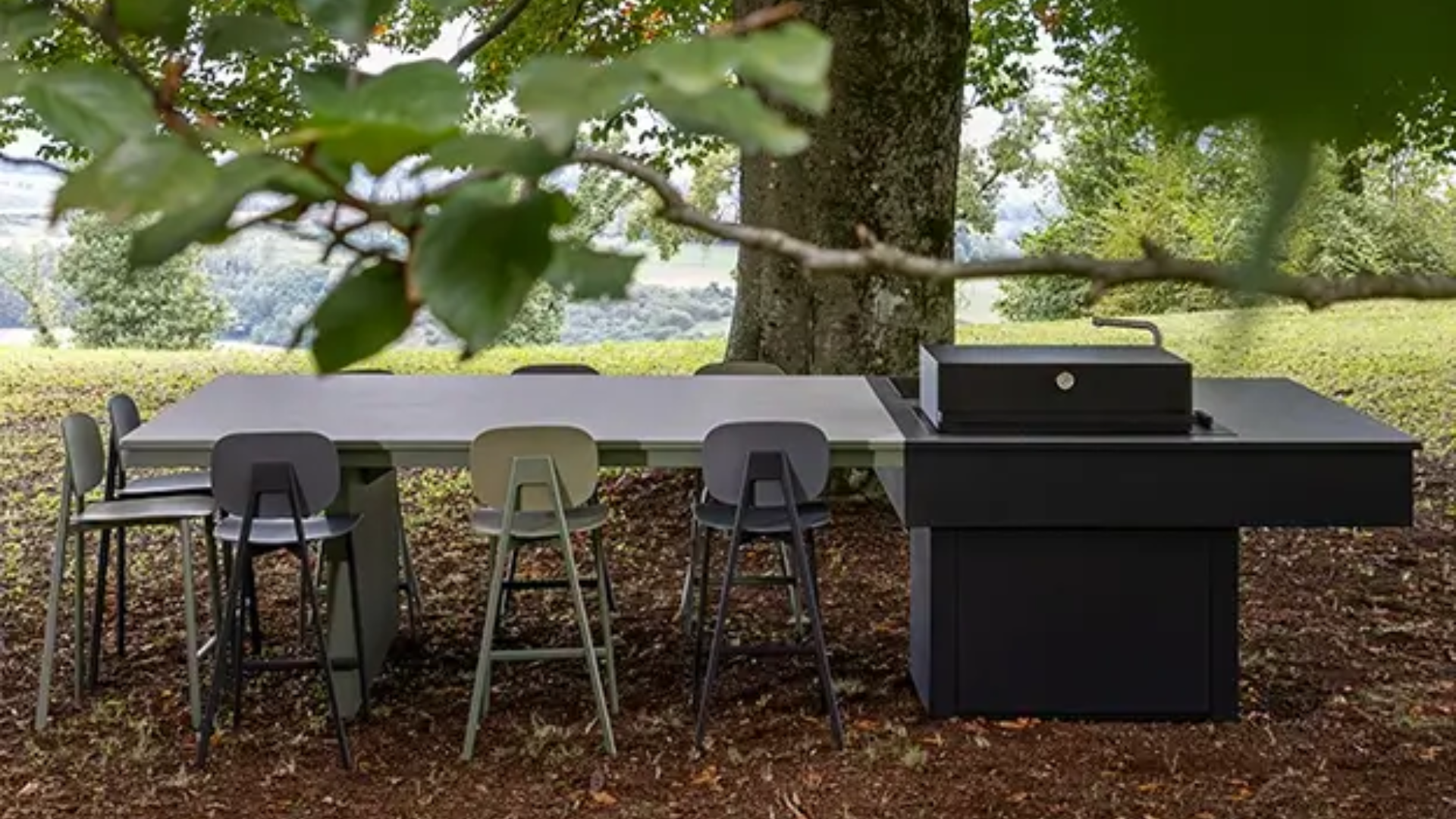 Cucina da esterno moderna di colore scuro con lungo tavolo e sedie di design disposta sotto la chioma di un albero in giardino.