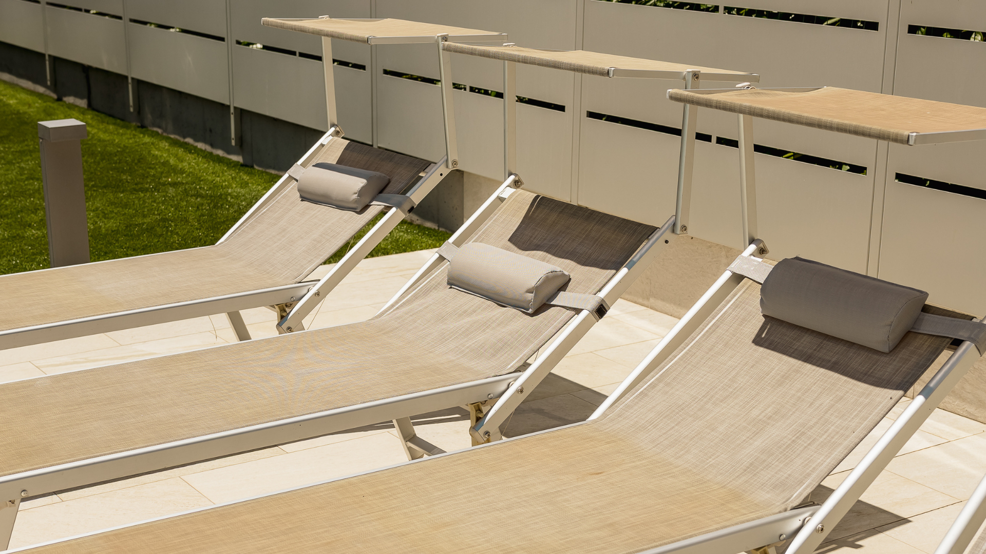 Area solarium con quattro lettini da giardino di design in tonalità neutre con parasole, disposti su pavimentazione in pietra.