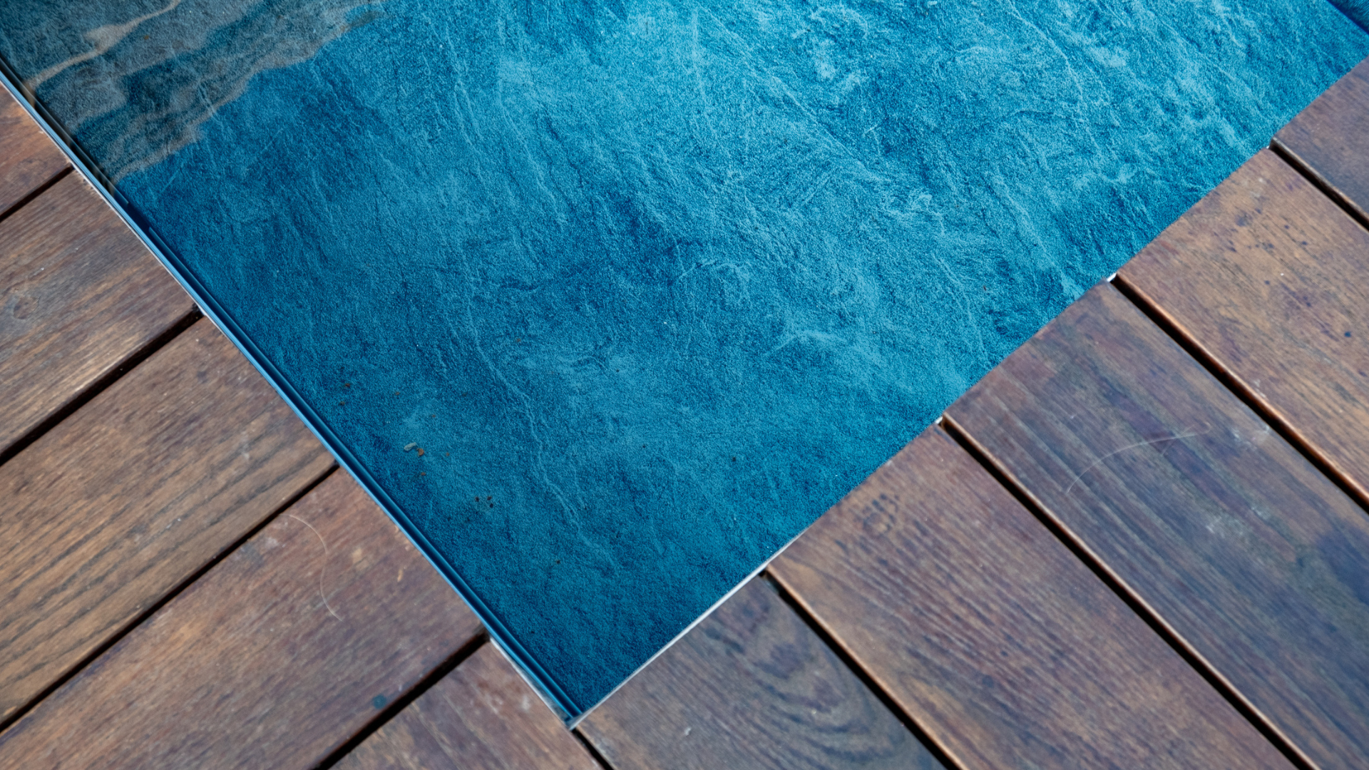 Dettaglio ravvicinato dell'incontro tra la finitura blu effetto pietra della piscina e il pavimento in parquet da esterno in legno.