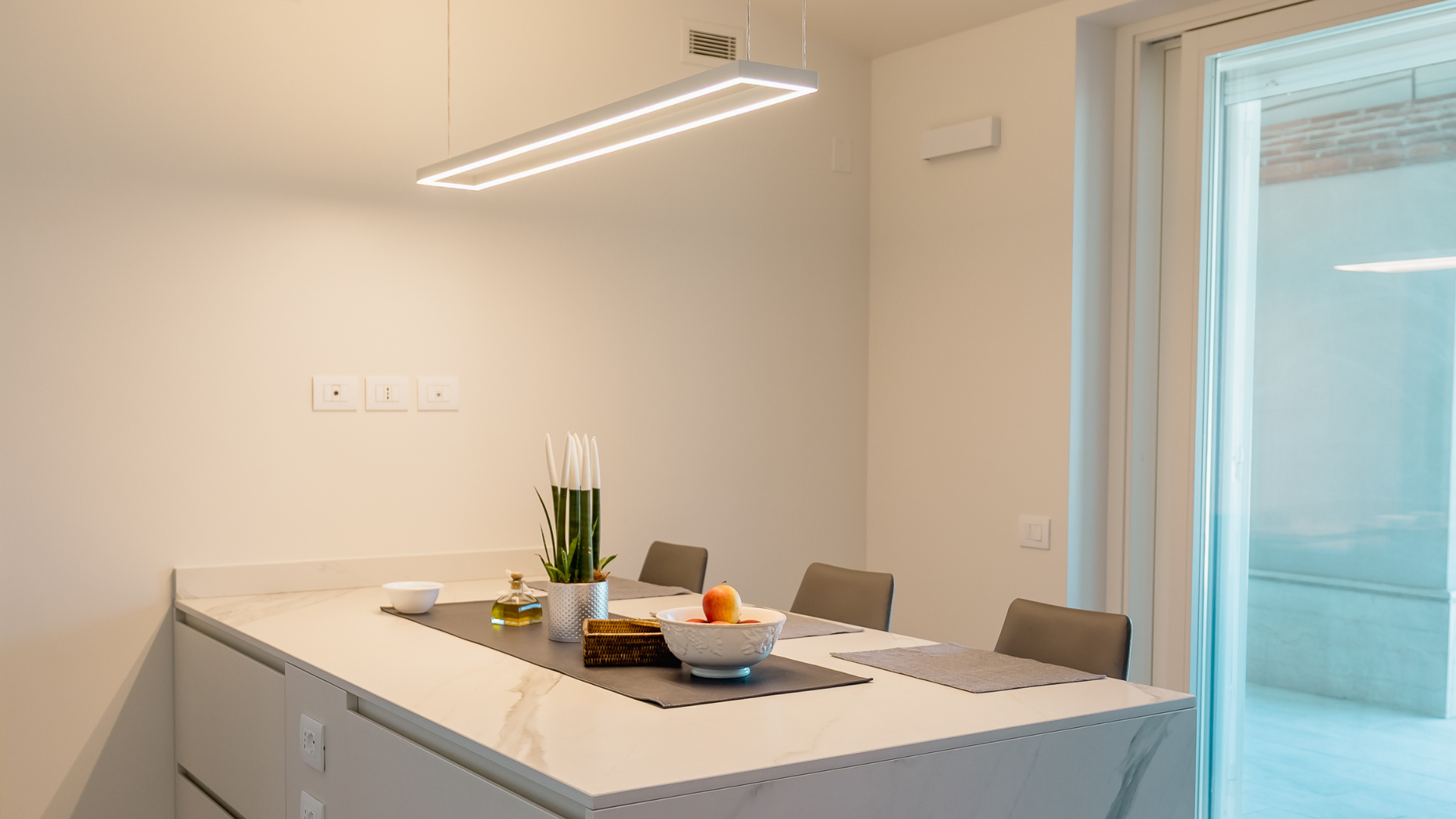 Isola da cucina in marmo chiaro con lampada a sospensione a LED e porta finestra, integrata con sistema di deumidificazione estiva e ricambio d'aria automatico.