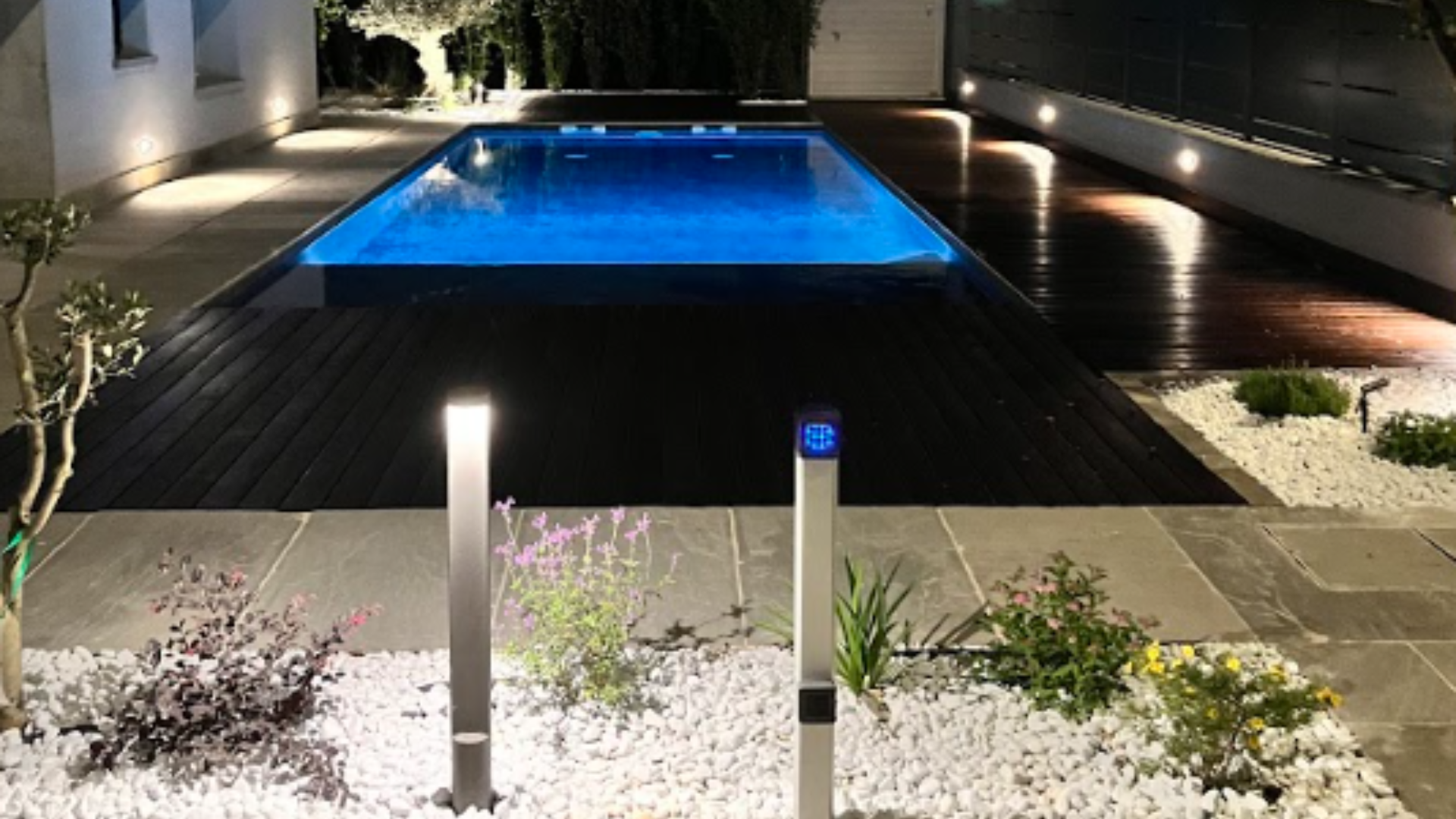Vista notturna di una piscina illuminata in un giardino moderno con pavimentazione in pietra, decking in legno e punti luce segnapasso.