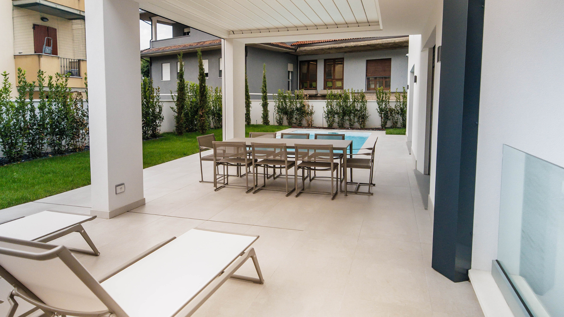 Patio esterno moderno con pergola bioclimatica, tavolo da pranzo outdoor, lettini prendisole e piscina interrata sullo sfondo.