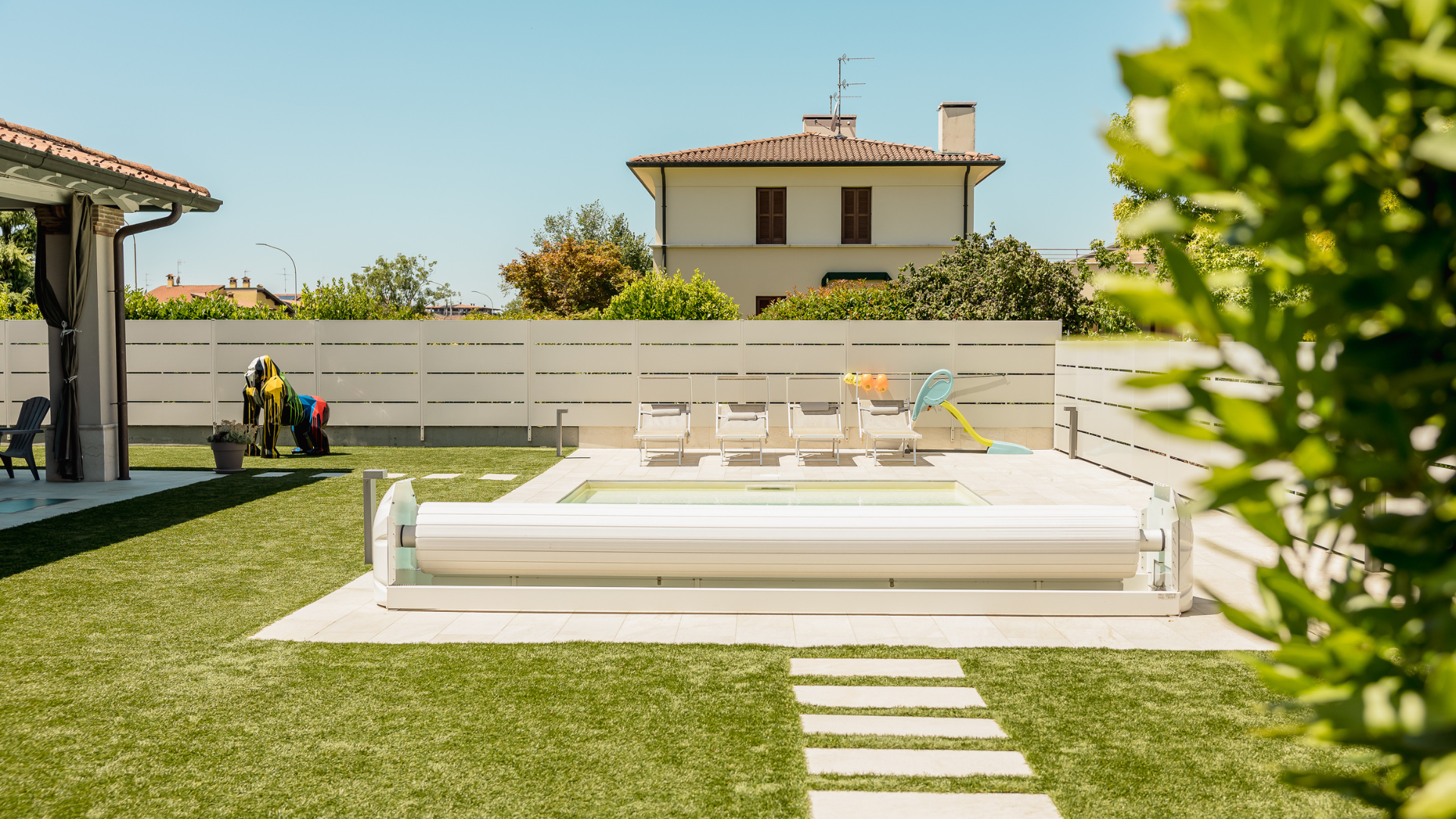 Zona solarium e piscina interrata dotata di copertura automatica a tapparella in un giardino moderno curato nei dettagli.