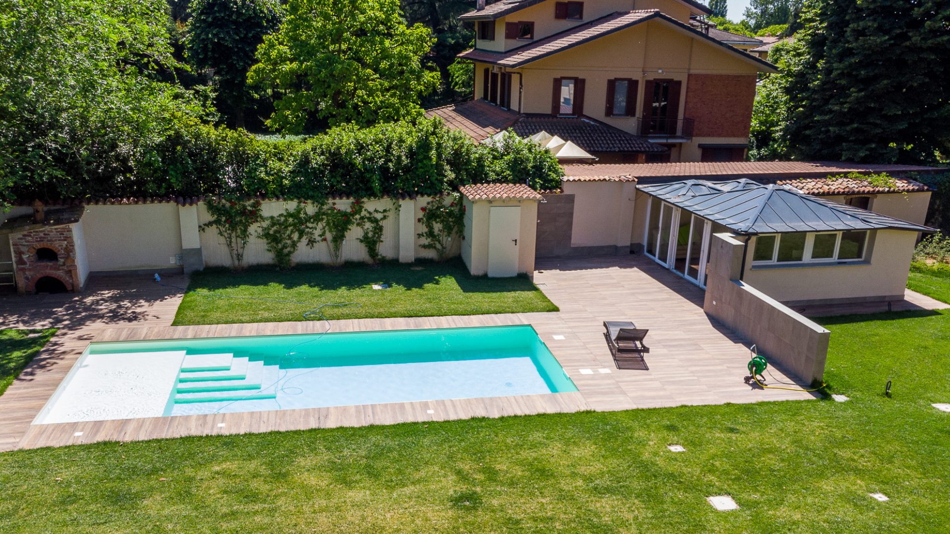 Vista prospettica di una piscina interrata con bordo in legno immersa nel verde di un giardino privato con prato all'inglese.
