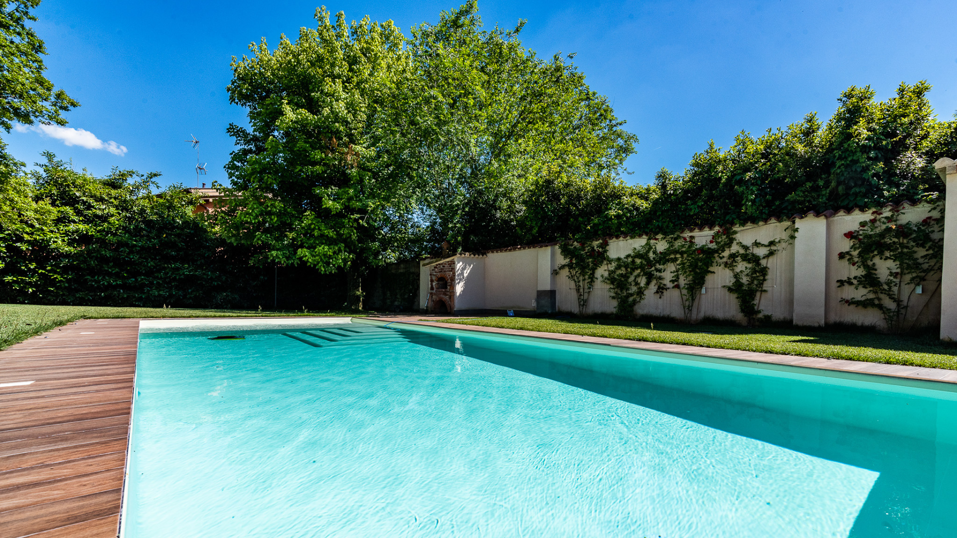Prospettiva laterale di una piscina interrata con zona solarium in piastrelle chiare, lettini prendisole e facciata di una villa residenziale sullo sfondo.