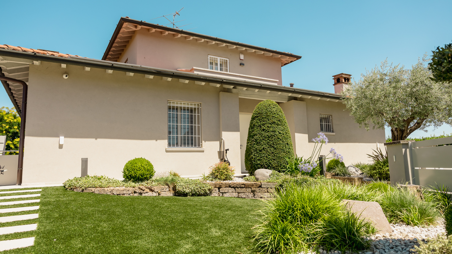 Facciata esterna di una villa residenziale ristrutturata con giardino curato, camminamento in pietra, prato all'inglese e aiuola decorativa con pianta di ulivo.