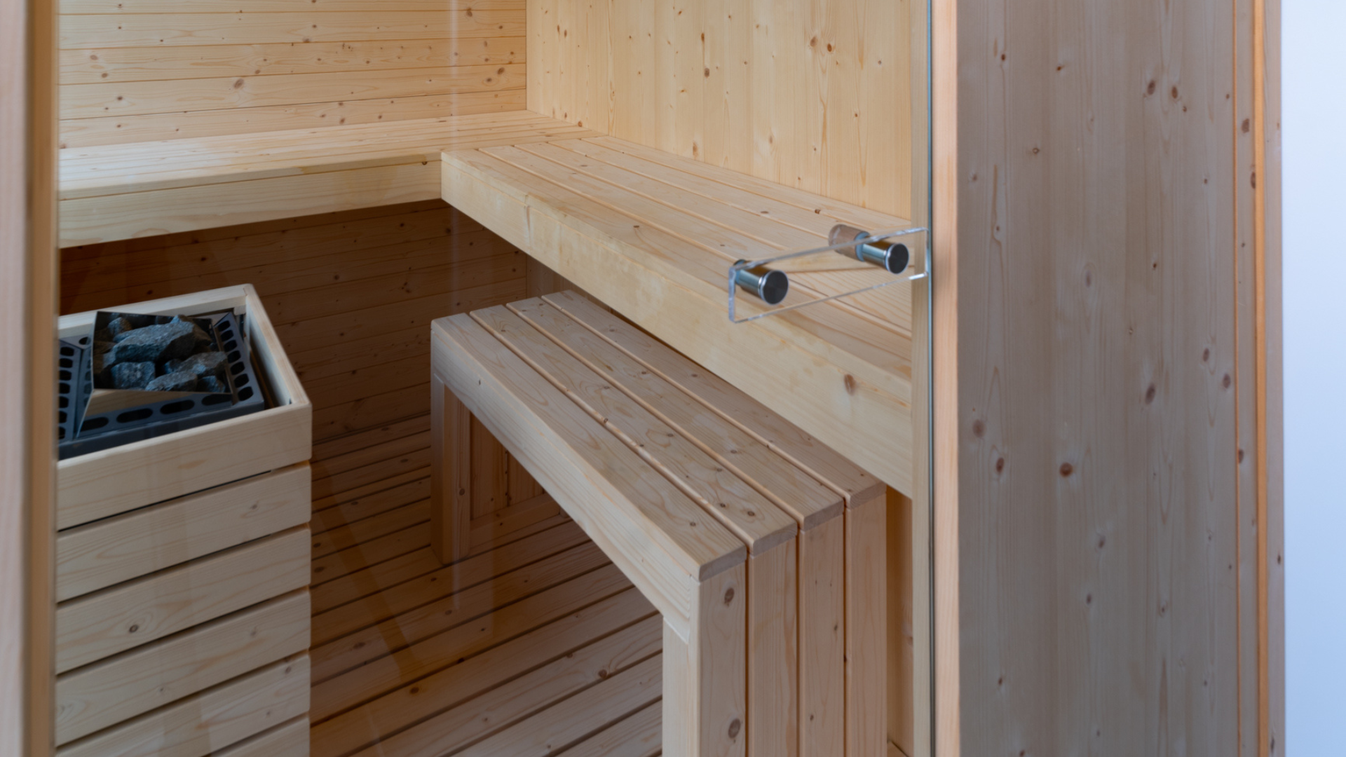 Interno di una sauna moderna in legno chiaro con panche sospese, luce LED d'ambiente e porta in vetro.