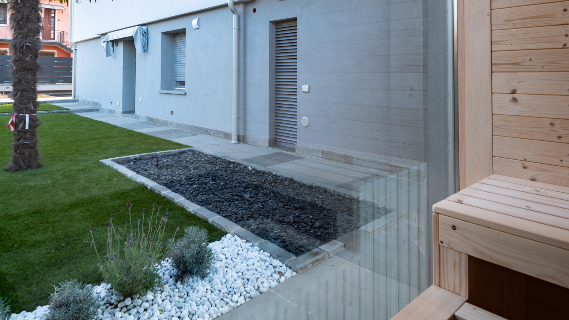 Vista dall'interno di una sauna in legno con vetrata affacciata sul giardino privato, prato verde e aiuola con ciottoli bianchi.