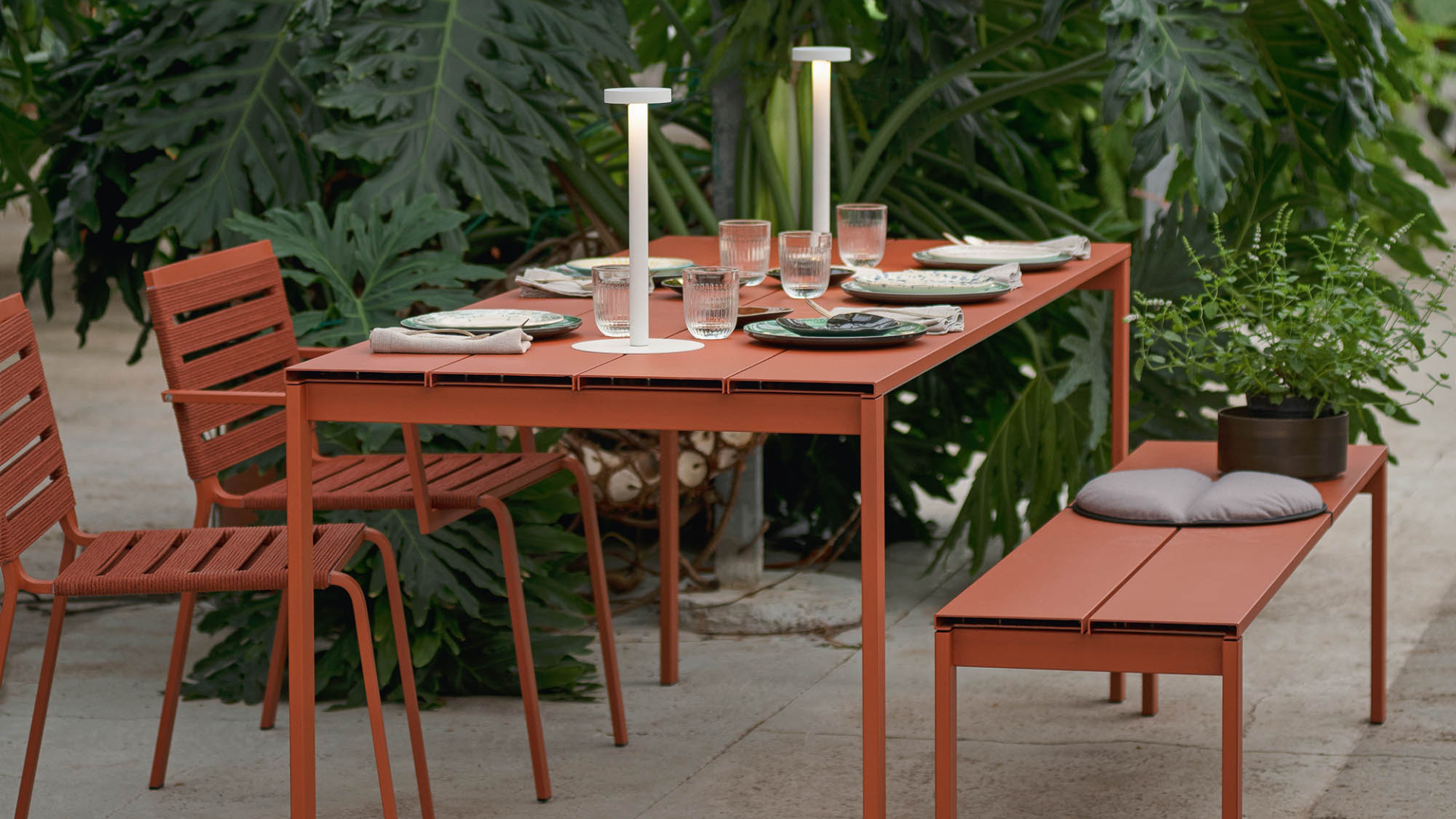 Tavolo da pranzo e panchina color terracotta disposti all'interno di una serra o veranda con piante tropicali e lampade da tavolo moderne.
