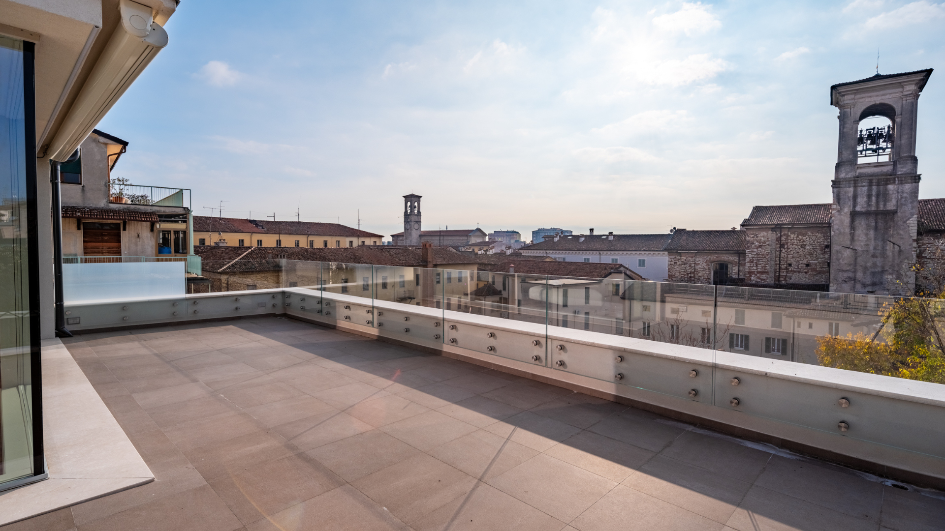 Ampia terrazza panoramica di un attico moderno con parapetto in vetro trasparente, pavimentazione outdoor e vista sui tetti del centro storico con campanile.