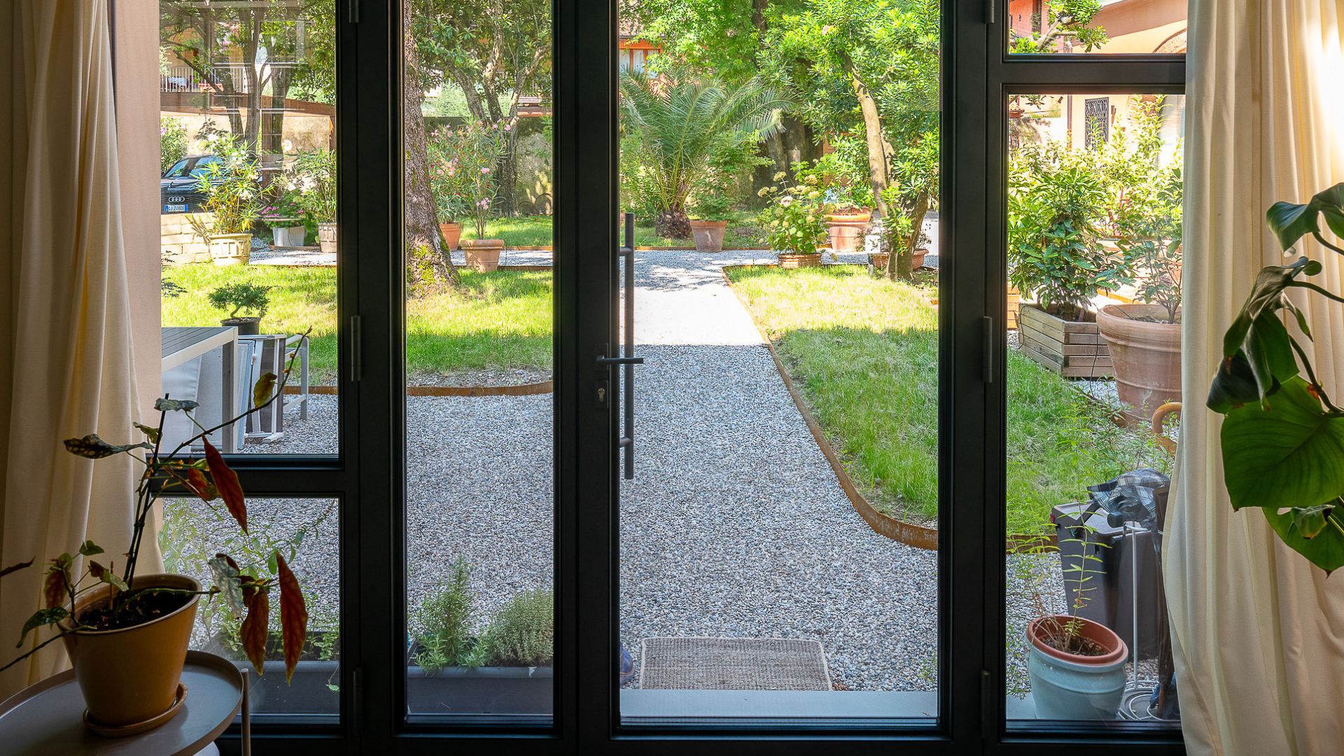 Vetrata ad arco con serramento scuro che incornicia la vista su un giardino privato verde con camminamento in ghiaia. 